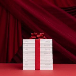 A neatly stacked collection of white books is tied together with a bold red ribbon and bow, set against a rich red draped fabric background. The titles on the book spines indicate they are religious texts, adding a reverent and celebratory ambiance.