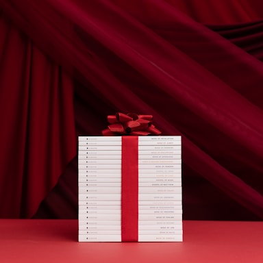 A neatly stacked collection of white books is tied together with a bold red ribbon and bow, set against a rich red draped fabric background. The titles on the book spines indicate they are religious texts, adding a reverent and celebratory ambiance.