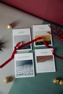 Four books are laid out on a table, each featuring a photographic cover and the names 'John,' 'Luke,' 'Matthew,' and 'Mark' printed above. A red ribbon weaves among the books, adding a festive touch. Decorative gold baubles and red berries are placed nearby, contributing to a holiday-themed arrangement.