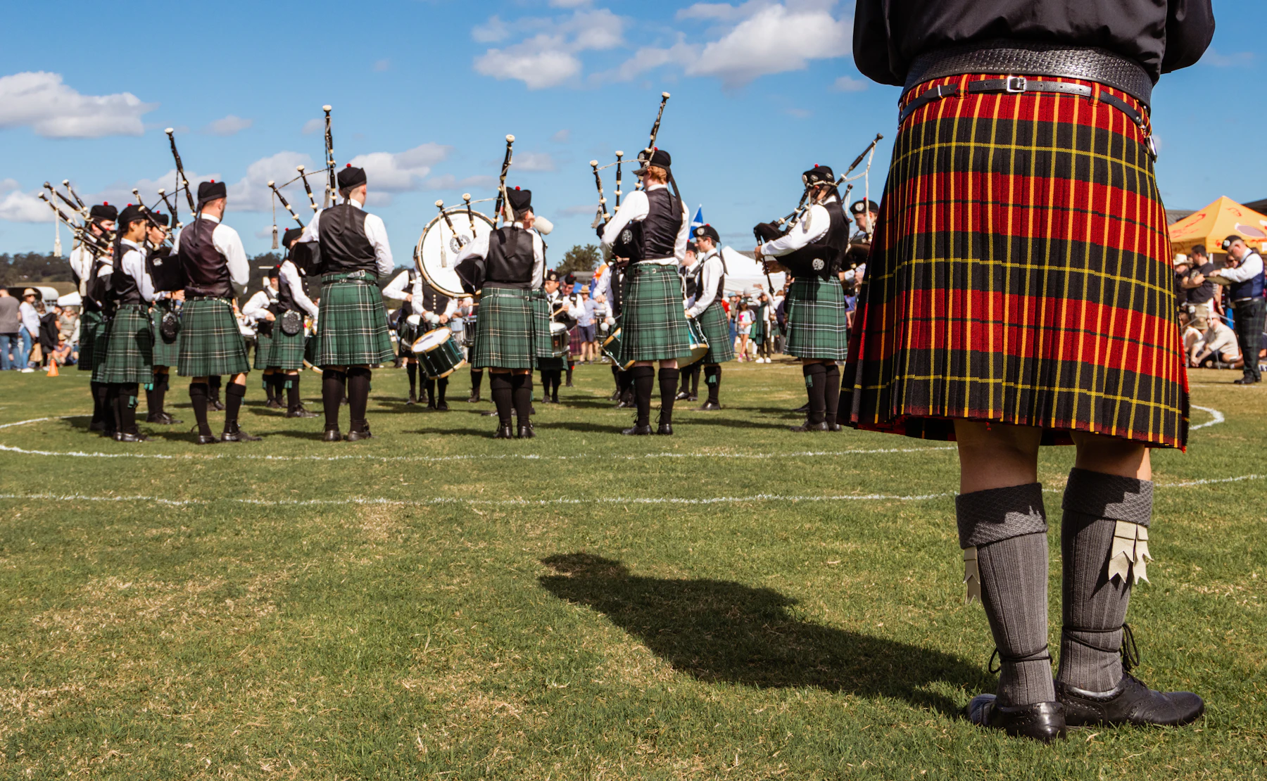 A pipe band in green kilts formed up on a grass games field, with a kilted figure in knee-high hose standing in the foreground