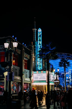 A vibrant, nighttime urban scene featuring a retro cinema with a neon sign reading 'PANTAGES'. The theater's marquee lists the names Zhang Yimou and Steven Spielberg. Bright streetlights illuminate the area, and groups of people walk along the bustling street, creating a lively atmosphere. Palm trees and Art Deco architecture enhance the scene's classic Hollywood vibe.