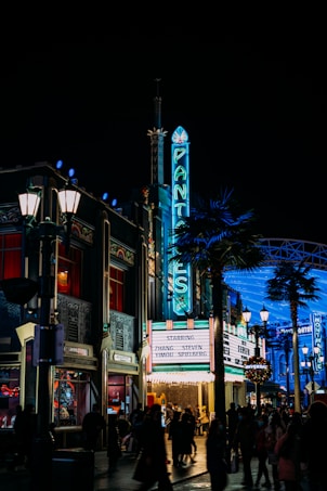 A vibrant, nighttime urban scene featuring a retro cinema with a neon sign reading 'PANTAGES'. The theater's marquee lists the names Zhang Yimou and Steven Spielberg. Bright streetlights illuminate the area, and groups of people walk along the bustling street, creating a lively atmosphere. Palm trees and Art Deco architecture enhance the scene's classic Hollywood vibe.