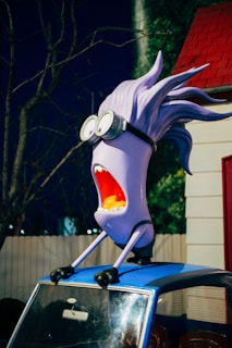 A cartoonish, purple character with wild hair and large goggles is mounted on top of a blue car. The character has a wide-open mouth, appearing to be yelling or surprised. The setting is outdoors in front of a structure with a red roof, and there are bare tree branches visible in the background.