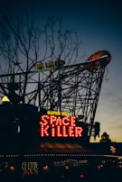 A neon sign with the words 'Super Silly Space Killer' is illuminated against a darkening sky. The sign is attached to a structure resembling a roller coaster, with decorative lights and an elaborate design visible in the background. Bare trees are silhouetted against the sky, adding an eerie atmosphere.