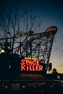 A neon sign with the words 'Super Silly Space Killer' is illuminated against a darkening sky. The sign is attached to a structure resembling a roller coaster, with decorative lights and an elaborate design visible in the background. Bare trees are silhouetted against the sky, adding an eerie atmosphere.