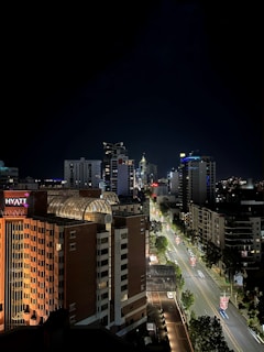 A nighttime cityscape featuring illuminated skyscrapers and buildings. The street in the foreground is lined with trees and adorned with hanging lights or banners. A prominent hotel building with the name 'Hyatt' is visible to the left.