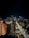 A nighttime cityscape featuring illuminated skyscrapers and buildings. The street in the foreground is lined with trees and adorned with hanging lights or banners. A prominent hotel building with the name 'Hyatt' is visible to the left.