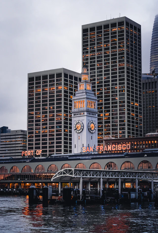 The Ferry Building's clock tower lit at dusk above its arched facade reading PORT OF SAN FRANCISCO, with skyscrapers behind.