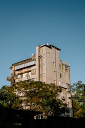 A multi-story residential building with a beige facade stands amidst greenery. The structure exhibits urban architecture with visible balconies and exterior pipework. Lush trees partially obscure the lower part of the building, while the clear, blue sky forms the backdrop.