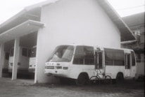 A black and white image of a parked bus with visible wear, next to a building. There is a bicycle leaning against the front of the bus, and another bus partially visible inside a garage on the left.