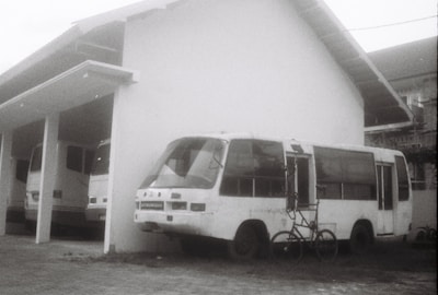 A black and white image of a parked bus with visible wear, next to a building. There is a bicycle leaning against the front of the bus, and another bus partially visible inside a garage on the left.