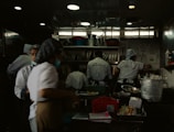 A busy commercial kitchen with several chefs in white uniforms and hats working diligently. The chefs appear to be preparing food amidst various cooking utensils and large pots. The environment is dimly lit with a focus on functionality.