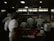 A busy commercial kitchen with several chefs in white uniforms and hats working diligently. The chefs appear to be preparing food amidst various cooking utensils and large pots. The environment is dimly lit with a focus on functionality.