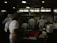 A busy commercial kitchen with several chefs in white uniforms and hats working diligently. The chefs appear to be preparing food amidst various cooking utensils and large pots. The environment is dimly lit with a focus on functionality.