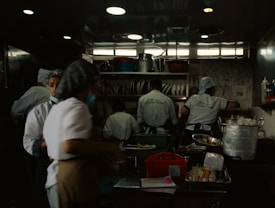 A busy commercial kitchen with several chefs in white uniforms and hats working diligently. The chefs appear to be preparing food amidst various cooking utensils and large pots. The environment is dimly lit with a focus on functionality.