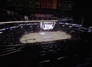 An indoor ice hockey arena with an illuminated scoreboard and a partially filled audience. The ice rink is visible with graphics projected onto the surface, and banners hang from the ceiling. Players are on the ice, and spectators are scattered throughout the stands.