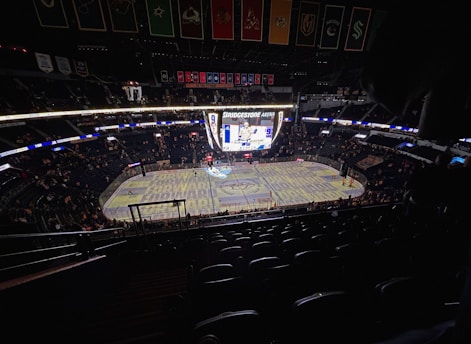An indoor ice hockey arena with an illuminated scoreboard and a partially filled audience. The ice rink is visible with graphics projected onto the surface, and banners hang from the ceiling. Players are on the ice, and spectators are scattered throughout the stands.