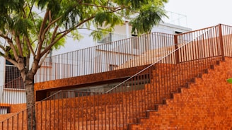 A modern structure made of red bricks features a staircase and railing, with a tree providing shade. The building has a minimalist design and incorporates natural elements.