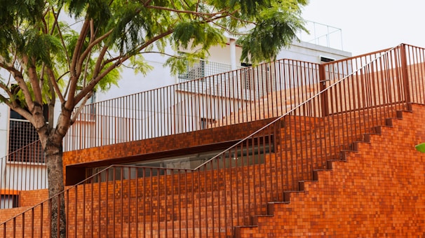 A modern structure made of red bricks features a staircase and railing, with a tree providing shade. The building has a minimalist design and incorporates natural elements.