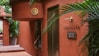The scene features the entrance of a spa with red-brown tiled walls. There is greenery in the form of potted plants, adding a touch of nature to the setting. A sign reads 'LOESS SPA' with additional information in smaller text. Stylish elements like the gold emblem and minimalist decor create an elegant atmosphere.