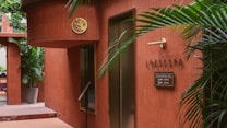The scene features the entrance of a spa with red-brown tiled walls. There is greenery in the form of potted plants, adding a touch of nature to the setting. A sign reads 'LOESS SPA' with additional information in smaller text. Stylish elements like the gold emblem and minimalist decor create an elegant atmosphere.