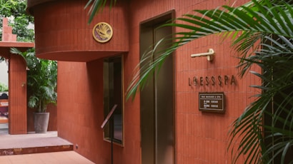 The scene features the entrance of a spa with red-brown tiled walls. There is greenery in the form of potted plants, adding a touch of nature to the setting. A sign reads 'LOESS SPA' with additional information in smaller text. Stylish elements like the gold emblem and minimalist decor create an elegant atmosphere.