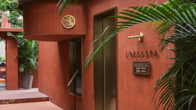 The scene features the entrance of a spa with red-brown tiled walls. There is greenery in the form of potted plants, adding a touch of nature to the setting. A sign reads 'LOESS SPA' with additional information in smaller text. Stylish elements like the gold emblem and minimalist decor create an elegant atmosphere.
