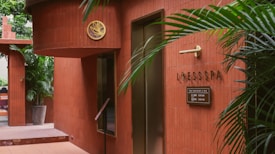 The scene features the entrance of a spa with red-brown tiled walls. There is greenery in the form of potted plants, adding a touch of nature to the setting. A sign reads 'LOESS SPA' with additional information in smaller text. Stylish elements like the gold emblem and minimalist decor create an elegant atmosphere.