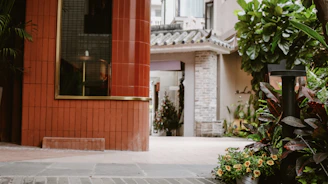 A peaceful courtyard area framed by lush greenery and flowering plants. A large red-tiled column is on one side, contrasting with a background of traditional architectural elements, including a brick wall and tiled roof. The setting seems serene and inviting, with a mix of modern and classic design.