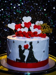 A beautifully decorated cake with a white base and red band, adorned with red and white heart-shaped decorations and small red roses on top. There is a silhouette of a couple, with the man kneeling, holding a heart in front of the woman. The word 'Happy Anniversary' is written in gold on top amidst the heart shapes. The background features glittering lights, creating a festive atmosphere.