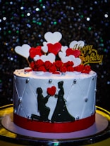A beautifully decorated cake with a white base and red band, adorned with red and white heart-shaped decorations and small red roses on top. There is a silhouette of a couple, with the man kneeling, holding a heart in front of the woman. The word 'Happy Anniversary' is written in gold on top amidst the heart shapes. The background features glittering lights, creating a festive atmosphere.