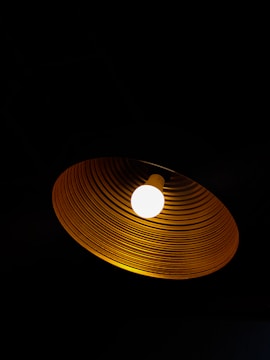 A single glowing light bulb is encased in a circular, ridged lampshade. The bulb emits a warm, bright light, contrasting sharply with the surrounding darkness.