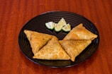 Three crispy golden-brown samosas are placed on a black round plate with a few wedges of lime. The plate is set against a rich wooden surface.