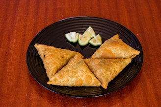 Three crispy golden-brown samosas are placed on a black round plate with a few wedges of lime. The plate is set against a rich wooden surface.