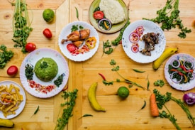A vibrant array of dishes displayed on a wooden table. There are plates with roasted chicken, rice, fried potatoes, and various sautéed vegetables. Fresh produce like bananas, tomatoes, chilies, a carrot, and leafy greens are scattered around. Each plate is artistically garnished with herbs and colorful sliced vegetables.