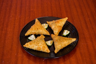 Four triangular pastries are arranged on a black plate, each accompanied by a small wedge of lime. The plate rests on a wooden surface.