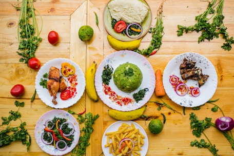 A colorful assortment of dishes featuring a variety of foods including green mashed potatoes, roasted chicken, fried plantains, grilled meat, and salad with onions and bell peppers. The dishes are creatively garnished and arranged on a wooden surface with scattered fresh herbs, tomatoes, carrots, and bananas.
