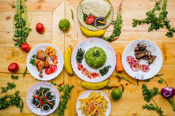 A colorful assortment of dishes featuring a variety of foods including green mashed potatoes, roasted chicken, fried plantains, grilled meat, and salad with onions and bell peppers. The dishes are creatively garnished and arranged on a wooden surface with scattered fresh herbs, tomatoes, carrots, and bananas.