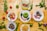 A colorful assortment of dishes featuring a variety of foods including green mashed potatoes, roasted chicken, fried plantains, grilled meat, and salad with onions and bell peppers. The dishes are creatively garnished and arranged on a wooden surface with scattered fresh herbs, tomatoes, carrots, and bananas.
