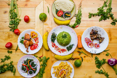 A colorful assortment of dishes featuring a variety of foods including green mashed potatoes, roasted chicken, fried plantains, grilled meat, and salad with onions and bell peppers. The dishes are creatively garnished and arranged on a wooden surface with scattered fresh herbs, tomatoes, carrots, and bananas.