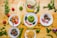 A colorful assortment of dishes featuring a variety of foods including green mashed potatoes, roasted chicken, fried plantains, grilled meat, and salad with onions and bell peppers. The dishes are creatively garnished and arranged on a wooden surface with scattered fresh herbs, tomatoes, carrots, and bananas.