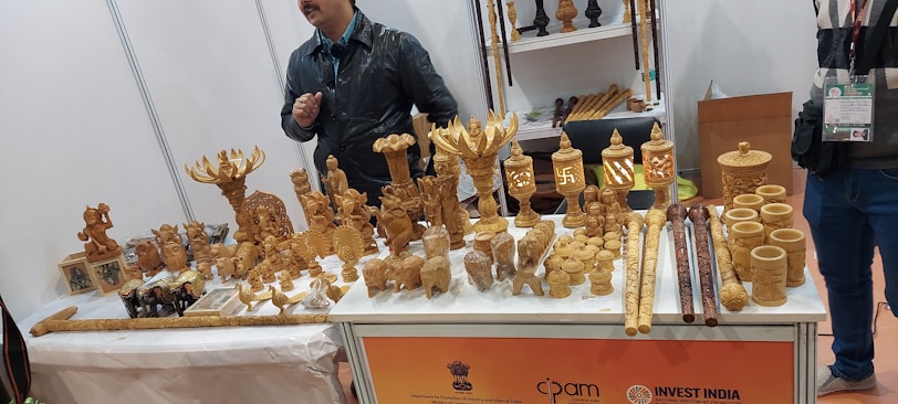 A display table at an exhibition showcasing a variety of intricately carved wooden handicrafts. The items include ornamental figures, small sculptures of deities and animals, cylindrical containers, and walking sticks. Two individuals stand behind the table, one wearing a black jacket.