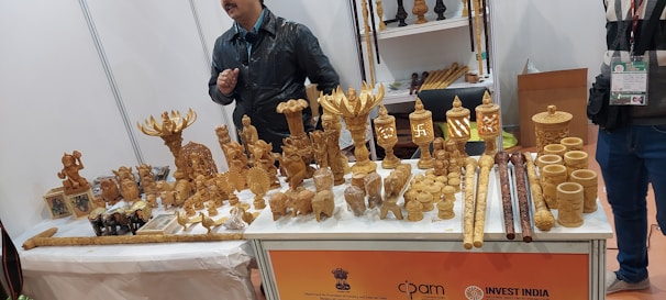 A display table at an exhibition showcasing a variety of intricately carved wooden handicrafts. The items include ornamental figures, small sculptures of deities and animals, cylindrical containers, and walking sticks. Two individuals stand behind the table, one wearing a black jacket.