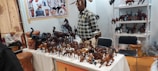 A vendor stands behind a table filled with various wooden animal sculptures, including elephants, horses, and camels. The background features display racks with more wooden animal carvings. There is a person sitting on the left, seemingly writing or drawing in a notebook. The setting appears to be a market or an exhibition.