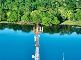 A tall communication tower stands prominently by a serene blue lake, surrounded by lush green forest. The water reflects the vibrant greenery of the trees, creating a peaceful and natural scene.