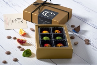 A box of colorful, artisanal chocolates with individual compartments is placed open on a white wooden surface. The chocolates are visually appealing with vibrant designs and speckled patterns. Beside the box is a closed gold gift box tied with a black ribbon. Scattered around are small chocolate pieces and slices of citrus fruits, adding a fresh contrast to the setup.