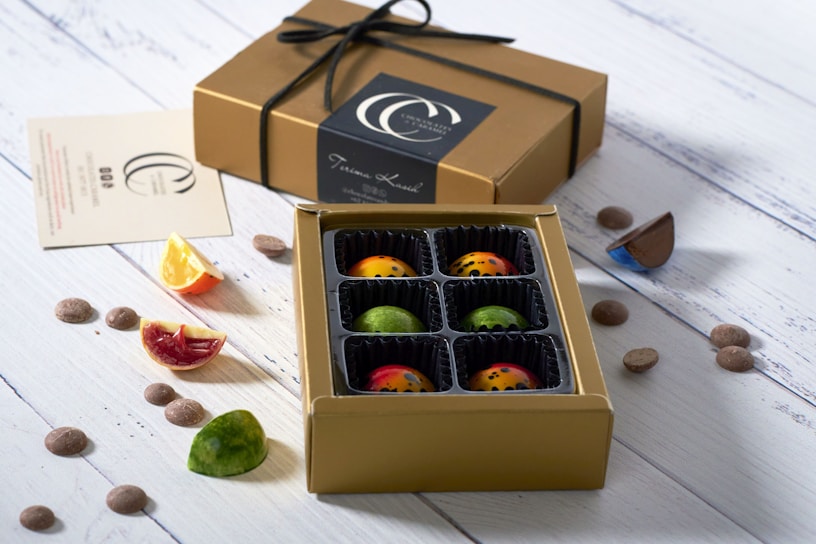 A box of colorful, artisanal chocolates with individual compartments is placed open on a white wooden surface. The chocolates are visually appealing with vibrant designs and speckled patterns. Beside the box is a closed gold gift box tied with a black ribbon. Scattered around are small chocolate pieces and slices of citrus fruits, adding a fresh contrast to the setup.