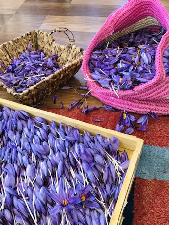 A collection of purple saffron flowers is spread out in a wooden tray and two woven baskets, one of which is pink. The flowers are vividly colored with bright orange stigmas. The surface they are on seems to be a wooden floor with a portion of a textured red and blue rug visible.