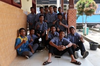 A group of young people wearing matching blue floral-patterned shirts and dark pants are gathered together, some sitting and some standing, in an outdoor setting next to a tiled wall. The individuals are displaying hand gestures like thumbs up and peace signs. The background includes a tree, a blue wall, and a tiled floor.