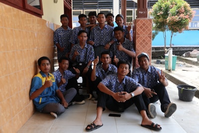 A group of young people wearing matching blue floral-patterned shirts and dark pants are gathered together, some sitting and some standing, in an outdoor setting next to a tiled wall. The individuals are displaying hand gestures like thumbs up and peace signs. The background includes a tree, a blue wall, and a tiled floor.
